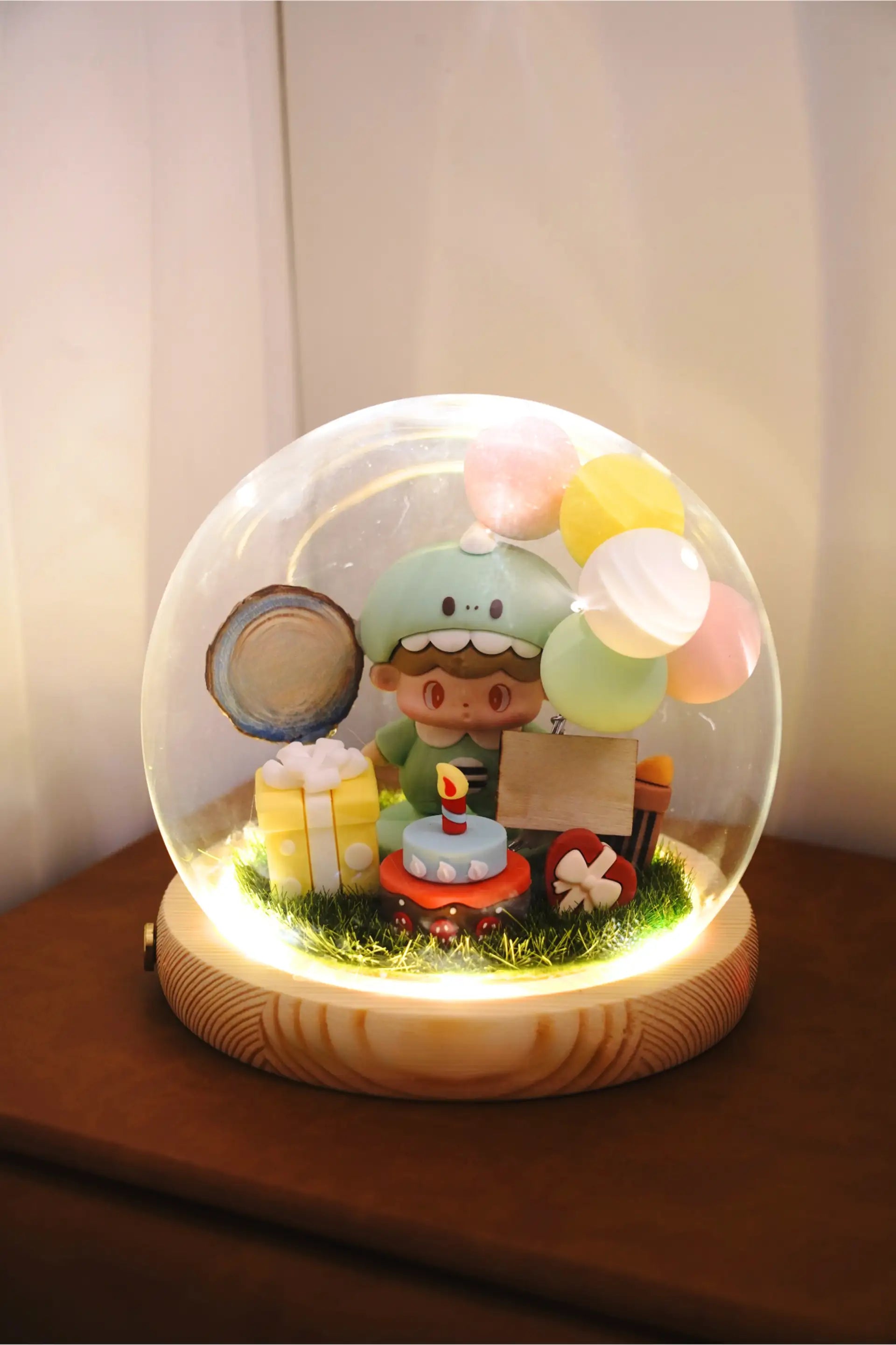 Baby birthday crystal ball featuring baby in dinosaur costume, birthday cake, gift boxes and balloons under round glass dome with LED lighting, blank wooden base for custom engraving