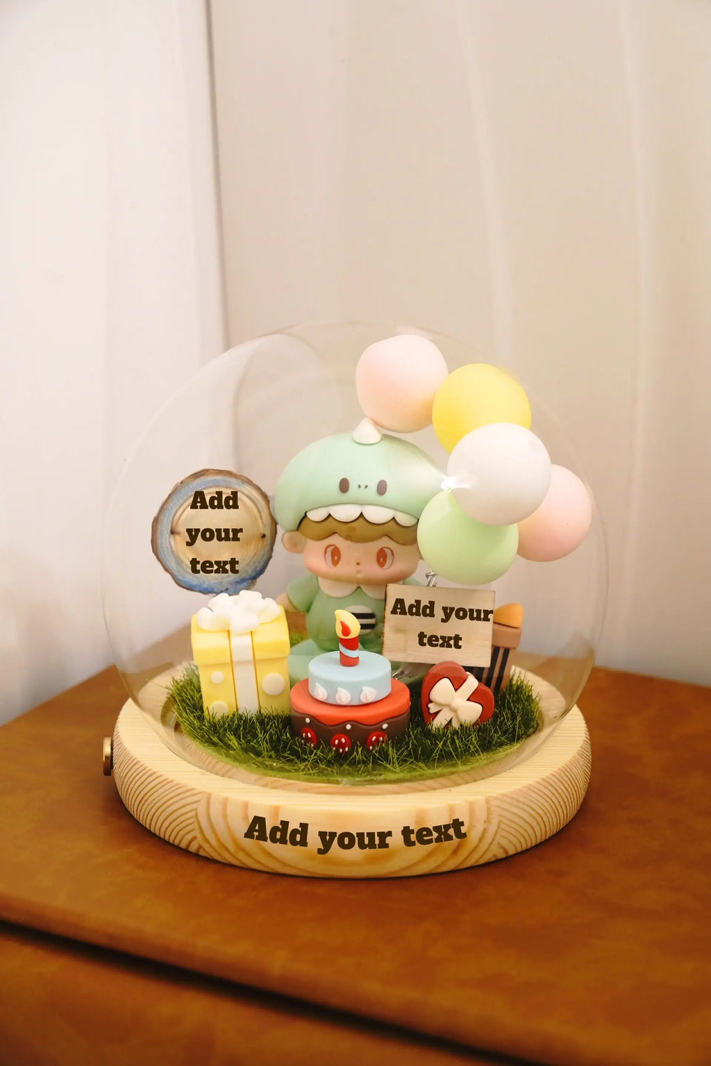Baby birthday crystal ball customisation guide showing engraving locations on wooden base with baby in dinosaur costume miniature, birthday cake and balloons inside dome